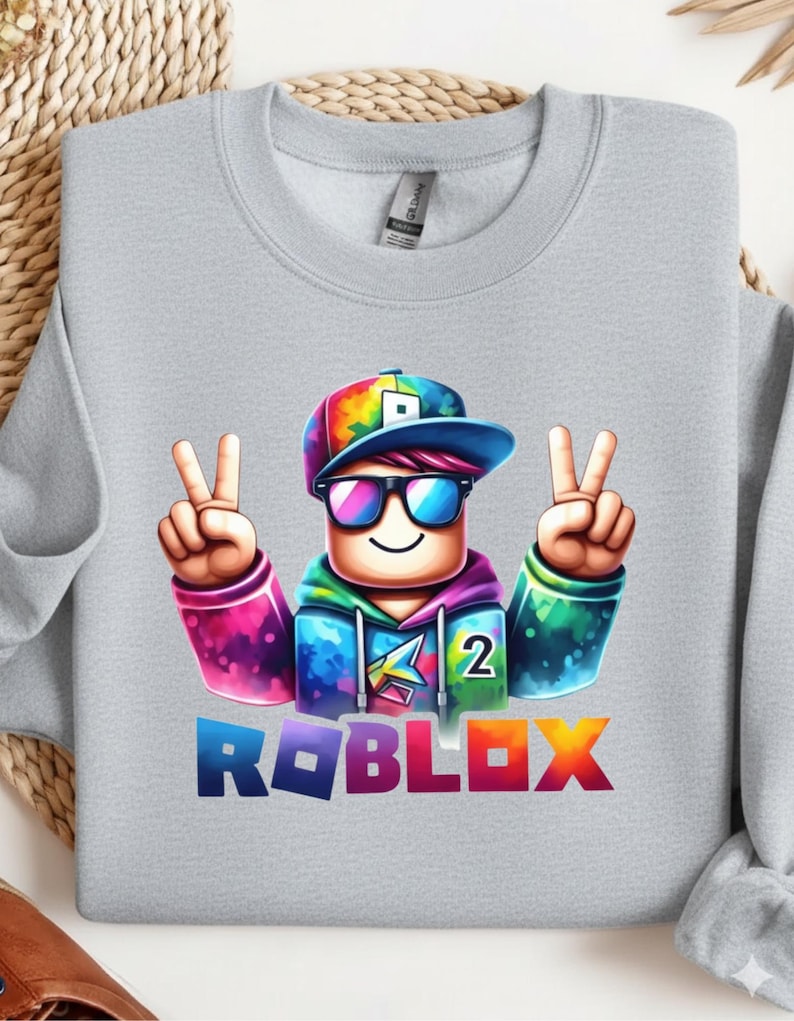 Puede incluir: Sudadera gris claro con un personaje de dibujos animados colorido con gafas de sol y un signo de la paz. El personaje lleva un gorro y una sudadera con capucha de arco&iacute;ris. La palabra "ROBLOX" est&aacute; impresa en la parte inferior.