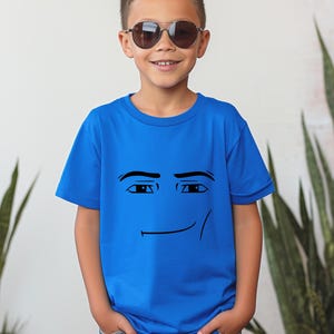 May include: A blue t-shirt featuring a smiling face graphic. The graphic is a simple black outline of eyes and a mouth. The shirt is worn by a person wearing sunglasses and blue jeans.