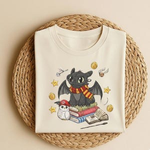 May include: Cream-colored t-shirt featuring a cartoon dragon wearing a scarf and glasses, sitting on books. The design includes an owl in a Mario hat, stars, coins, and a golden snitch. A whimsical design.