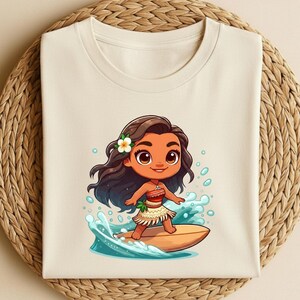 May include: Cream-colored t-shirt featuring a cartoon illustration of a girl surfing. The girl has brown hair, a flower, and a skirt. She is riding a surfboard on a wave. The shirt is folded on a woven, circular surface.