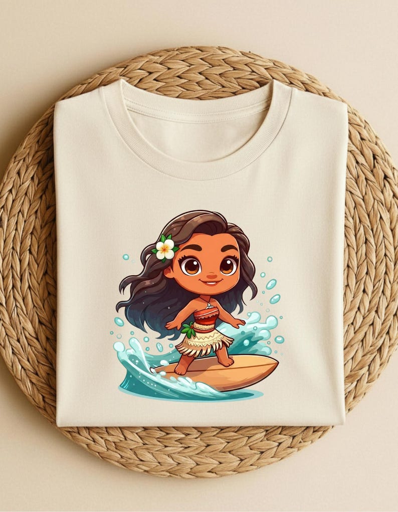 May include: Cream-colored t-shirt featuring a cartoon illustration of a girl surfing. The girl has brown hair, a flower, and a skirt. She is riding a surfboard on a wave. The shirt is folded on a woven, circular surface.