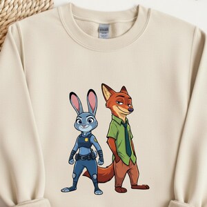 以下が含まれることがあります： 『ズートピア』のジュディ・ホップスとニック・ワイルドのグラフィックがプリントされたベージュのクルーネック・スウェットシャツ。ジュディは青い警察の制服を着ており、ニックは緑のシャツと茶色のズボンを着用しています。デザインはスウェットシャツの前面中央に配置されています。