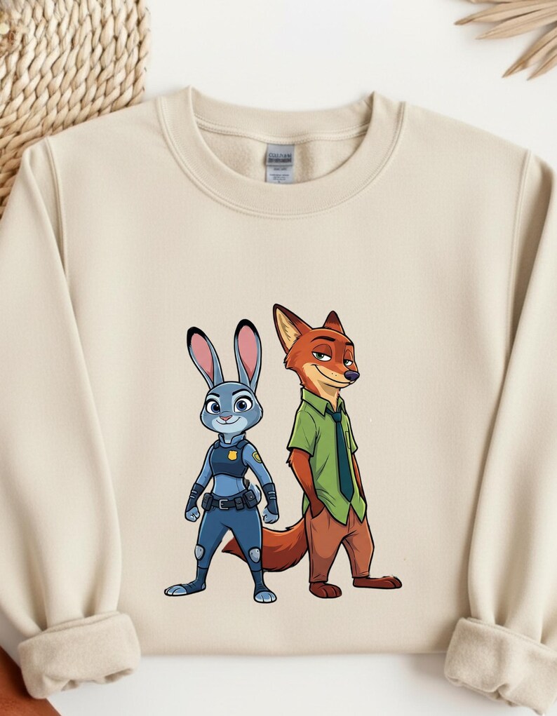 以下が含まれることがあります： 『ズートピア』のジュディ・ホップスとニック・ワイルドのグラフィックがプリントされたベージュのクルーネック・スウェットシャツ。ジュディは青い警察の制服を着ており、ニックは緑のシャツと茶色のズボンを着用しています。デザインはスウェットシャツの前面中央に配置されています。