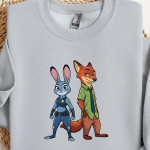 以下が含まれることがあります： ズートピアのキャラクター、ジュディ・ホップスとニック・ワイルドが描かれたライトグレーのスウェットシャツ。青いウサギのジュディは警察の制服を着ています。キツネのニックは緑色のシャツと茶色のズボンを着ています。スウェットシャツはクルーネックで長袖です。