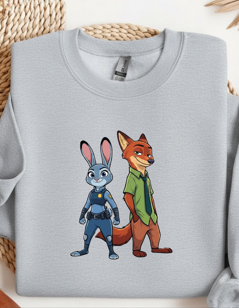 以下が含まれることがあります： ズートピアのキャラクター、ジュディ・ホップスとニック・ワイルドが描かれたライトグレーのスウェットシャツ。青いウサギのジュディは警察の制服を着ています。キツネのニックは緑色のシャツと茶色のズボンを着ています。スウェットシャツはクルーネックで長袖です。