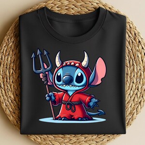 May include: Black t-shirt featuring a cartoon character in a red devil costume, holding a trident. The character is blue with large eyes and pink ears. The shirt is folded on a woven, circular surface.