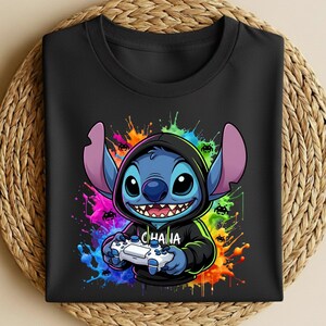 May include: Black t-shirt featuring a cartoon character with blue fur, large ears, and a hoodie, holding a video game controller. The design includes colorful paint splatters and the word "OHANA" on the hoodie.