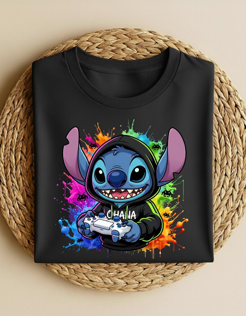 May include: Black t-shirt featuring a cartoon character with blue fur, large ears, and a hoodie, holding a video game controller. The design includes colorful paint splatters and the word "OHANA" on the hoodie.