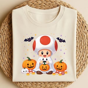 May include: A cream-colored t-shirt featuring a graphic of a mushroom character wearing a white ghost costume. The character is surrounded by three jack-o'-lantern pumpkins, small mushrooms, candy corn, and two black bats against a background of stars.