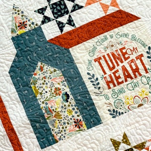 May include: A white quilt with a floral design and the text "Come Thou Fount of Every Blessing Tune My Heart To Sing Thy Grace". The quilt is made with a variety of fabrics, including a teal blue fabric with a floral pattern, a red fabric with a floral pattern, and a green fabric with a floral pattern.