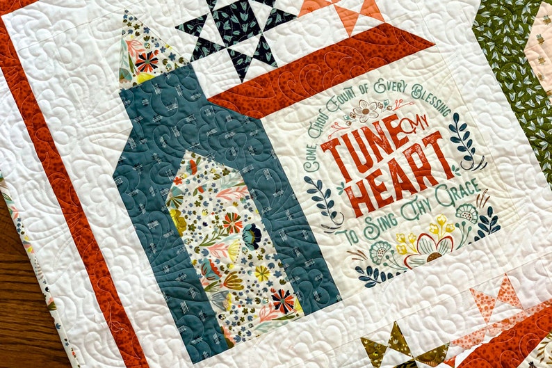 May include: A white quilt with a floral design and the text "Come Thou Fount of Every Blessing Tune My Heart To Sing Thy Grace". The quilt is made with a variety of fabrics, including a teal blue fabric with a floral pattern, a red fabric with a floral pattern, and a green fabric with a floral pattern.