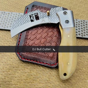 May include: A curved blade knife with a light-colored handle and a silver blade, resting on a brown leather sheath with red stitching. The blade has a decorative pattern and the text "DJ Bull Cutter".