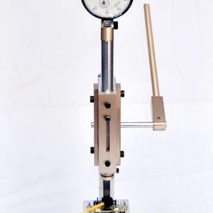 Op de afbeelding: Een zilveren en zwarte wijzerplaatmeter met een wit gezicht en zwarte markeringen. De meter is bevestigd aan een zilveren metalen staaf met een zwarte handgreep. De staaf is bevestigd aan een zilveren metalen basis met een zwarte handgreep. De meter wordt gebruikt om de dikte van objecten te meten.
