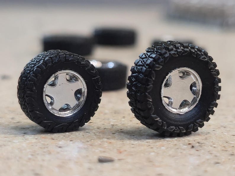 Puede incluir: Dos neum&aacute;ticos todoterreno en miniatura negros con llantas plateadas. Los neum&aacute;ticos tienen un dibujo profundo para un mejor agarre. Varios otros neum&aacute;ticos est&aacute;n ligeramente desenfocados en el fondo, lo que sugiere un juego.