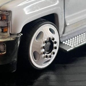 May include: Close-up of a white truck with a chrome bumper and a silver wheel. The wheel has a black tire and a silver rim with black lug nuts. A silver step is visible.