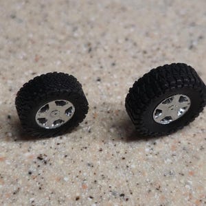 Pode incluir: S&atilde;o exibidos dois pneus pretos em miniatura para todo-o-terreno com aros prateados. Os pneus t&ecirc;m um padr&atilde;o de piso detalhado e os aros apresentam um design de cinco raios. Os pneus s&atilde;o provavelmente para um ve&iacute;culo modelo.