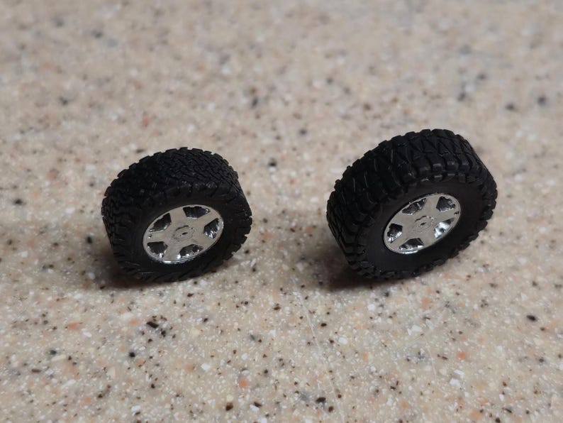 Pode incluir: S&atilde;o exibidos dois pneus pretos em miniatura para todo-o-terreno com aros prateados. Os pneus t&ecirc;m um padr&atilde;o de piso detalhado e os aros apresentam um design de cinco raios. Os pneus s&atilde;o provavelmente para um ve&iacute;culo modelo.