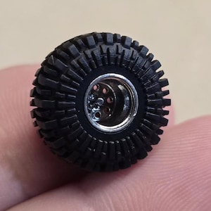 May include: A black, miniature off-road tire with a detailed tread pattern. The tire has a silver-colored wheel center with multiple holes. The tire is designed for a small-scale model vehicle.