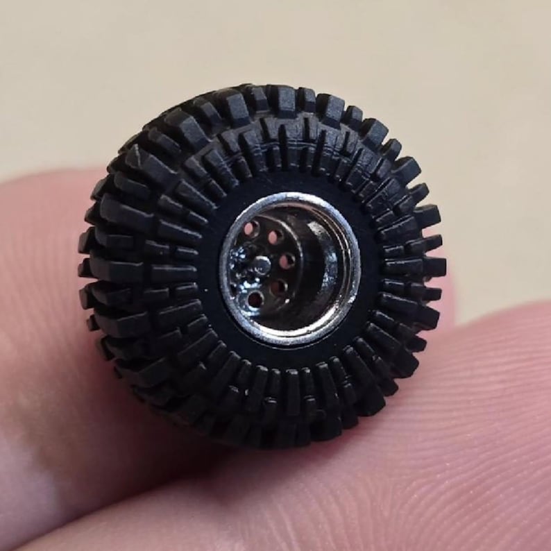 May include: A black, miniature off-road tire with a detailed tread pattern. The tire has a silver-colored wheel center with multiple holes. The tire is designed for a small-scale model vehicle.
