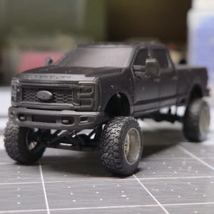 May include: A matte black model pickup truck with large off-road tires. The truck has a detailed front grille and the words "SUPER DUTY" are visible above the grille. The truck is sitting on a gray surface.