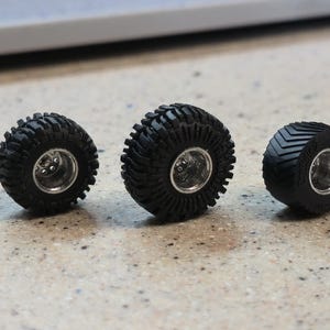 Pode incluir: Três pneus pretos em miniatura com aros prateados são exibidos em uma superfície salpicada. Os pneus têm diferentes padrões de banda de rodagem, incluindo um design com saliências e um padrão em zigue-zague. Estes são provavelmente pneus de modelos de carros ou caminhões.