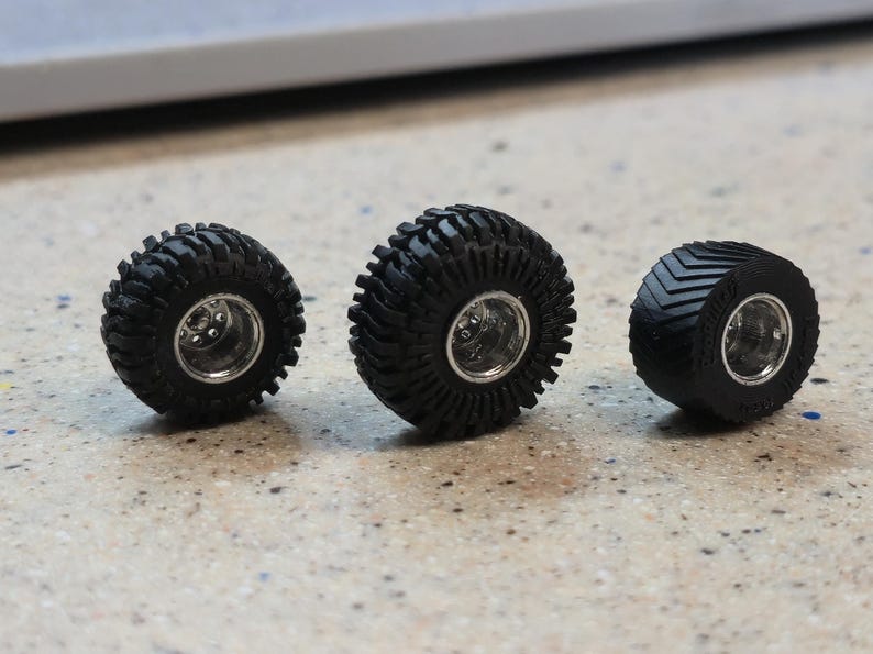 May include: Three black miniature tires with silver rims are displayed on a speckled surface. The tires have different tread patterns, including a knobby design and a chevron pattern. These are likely model car or truck tires.
