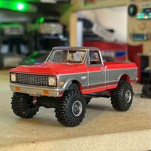 May include: A red and gray model pickup truck with large black tires. The truck has a silver roof and a detailed front grille. The model is a miniature replica of a classic truck.