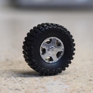 Pode incluir: Um pneu preto off-road com uma roda prateada de cinco raios. O pneu tem um padr&atilde;o de banda de rodagem profundo para maior ader&ecirc;ncia. A roda &eacute; de cor prateada brilhante e met&aacute;lica. Este &eacute; um modelo em miniatura de uma roda de ve&iacute;culo.