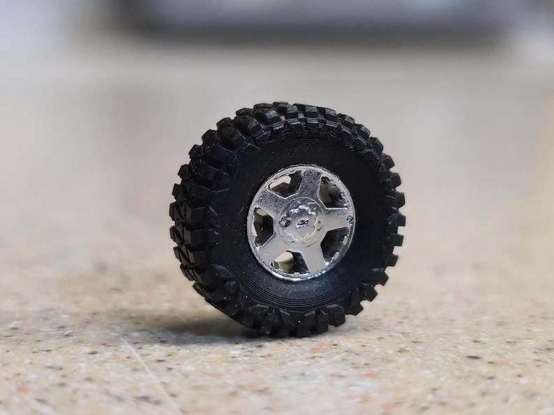 Pode incluir: Um pneu preto off-road com uma roda prateada de cinco raios. O pneu tem um padr&atilde;o de banda de rodagem profundo para maior ader&ecirc;ncia. A roda &eacute; de cor prateada brilhante e met&aacute;lica. Este &eacute; um modelo em miniatura de uma roda de ve&iacute;culo.