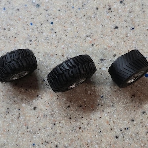 May include: Three black miniature tires with silver rims are arranged on a speckled surface. The tires have different tread patterns, suggesting a variety of uses. These small tires are likely for model vehicles or similar projects.
