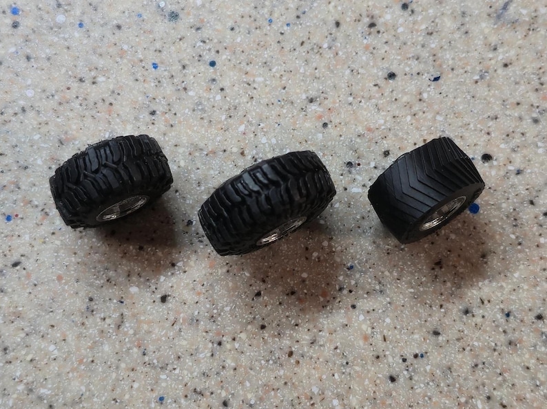 May include: Three black miniature tires with silver rims are arranged on a speckled surface. The tires have different tread patterns, suggesting a variety of uses. These small tires are likely for model vehicles or similar projects.