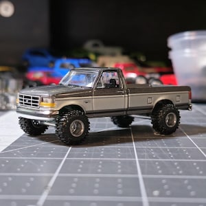 May include: A detailed model of a two-tone pickup truck with large, black off-road tires. The truck is primarily silver with a darker gray top and is set against a blurred background. The model has a detailed front grille and headlights.
