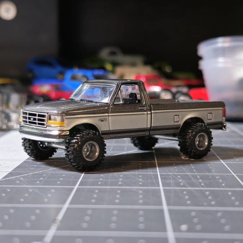 May include: A detailed model of a two-tone pickup truck with large, black off-road tires. The truck is primarily silver with a darker gray top and is set against a blurred background. The model has a detailed front grille and headlights.
