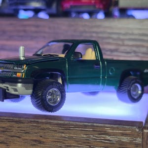 May include: A dark green model pickup truck with large off-road tires. The truck has a silver front bumper and a chrome exhaust stack. The interior is tan. The truck is displayed on a shelf with a blue light.