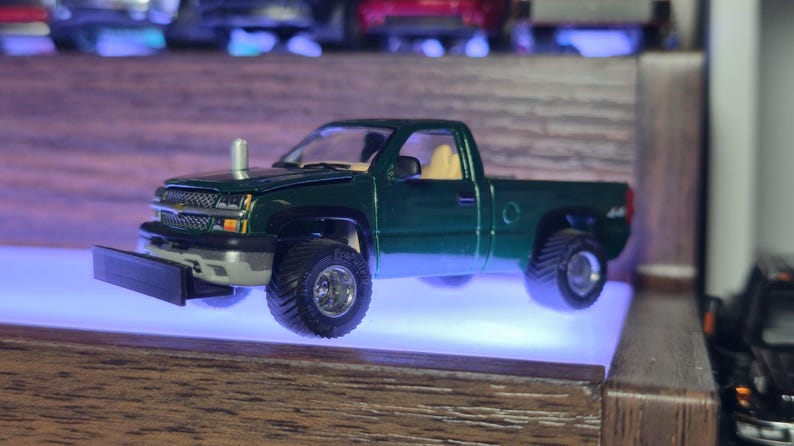 May include: A dark green model pickup truck with large off-road tires. The truck has a silver front bumper and a chrome exhaust stack. The interior is tan. The truck is displayed on a shelf with a blue light.