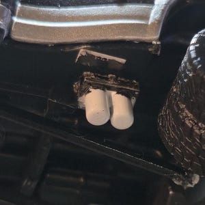 May include: Close-up of a model car's engine detail. The image shows a black chassis with a silver body panel. Two white cylindrical exhaust pipes are visible, along with a black engine component and a black tire.