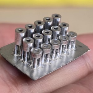 May include: A cluster of small, silver-colored electronic components arranged on a rectangular metal base. The components have cylindrical bodies with a hole in the center and multiple pins extending from the base. The image is a close-up shot.