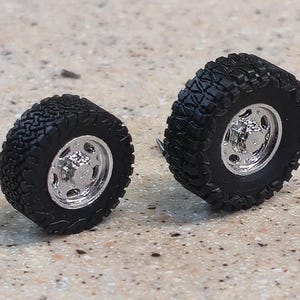 May include: Two miniature black off-road tires with chrome rims. The tires have a deep tread pattern, and the rims have a multi-hole design. The tires are likely for a model vehicle or a similar project.