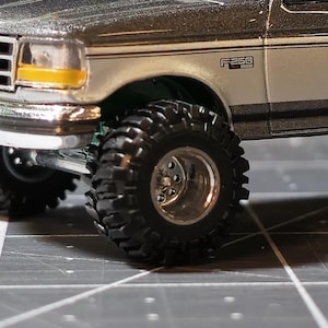 May include: Close-up of a silver and gray model truck with large, black off-road tires. The truck has a yellow headlight and the text "F250" on the side. The tires have a diameter of approximately 3 inches.