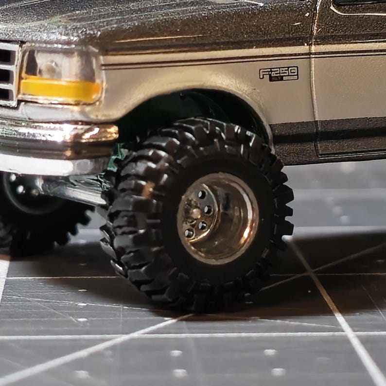 May include: Close-up of a silver and gray model truck with large, black off-road tires. The truck has a yellow headlight and the text "F250" on the side. The tires have a diameter of approximately 3 inches.