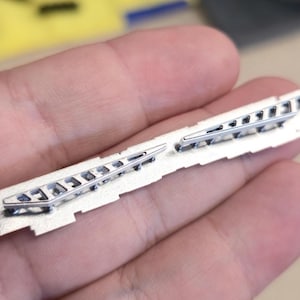 May include: Two silver-colored metal hair clips with a geometric design are displayed. The clips are attached to a light beige rectangular base. The clips have a series of small, rectangular cutouts.