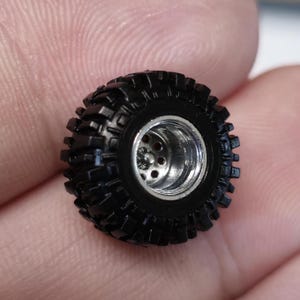 May include: Close-up of a small, black, off-road tire with a silver rim. The tire has a deep tread pattern for enhanced grip. The rim features multiple holes. This miniature tire is likely for a model vehicle.