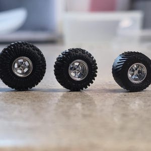 Pode incluir: Três pneus off-road pretos em miniatura com aros prateados estão dispostos em fila. Os pneus têm um padrão de banda de rodagem profundo para maior aderência. Os aros têm um design de cinco raios. Estes pneus pequenos são provavelmente para um veículo modelo.