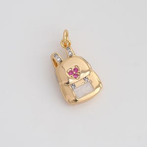 May include: A gold-colored backpack charm with a heart-shaped design made of small pink gems. The charm has a white rectangular base and is adorned with small clear gems on the straps. The charm is designed to be used as a pendant.