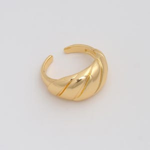 Puede incluir: Un anillo dorado con un diseño en forma de croissant. El anillo tiene una forma curva con crestas diagonales y un diseño abierto. El anillo está hecho de un metal brillante y reflectante.