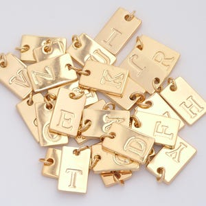 May include: A collection of gold-colored rectangular charms, each engraved with a different letter of the alphabet. The charms have a small loop at the top for attaching to a chain or bracelet. The letters are in a simple, sans-serif font.