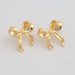 May include: A pair of gold-colored bow-shaped earrings. The earrings have a glossy finish and a detailed design, with the bow's loops and tails clearly defined. The earrings are presented against a plain, light background.