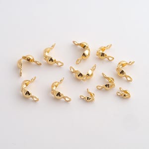 May include: A collection of gold-colored jewelry findings, including curved end caps and connectors. The findings have a rounded shape with small loops on each end for attaching to other jewelry components. The pieces are scattered on a white surface.