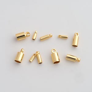 May include: A collection of gold-colored metal crimp end findings for jewelry making. The findings are cylindrical with a loop at one end for attaching to a chain or cord. The findings are in various sizes.