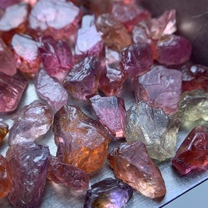 Puede incluir: Una colección de piedras preciosas en bruto y sin pulir en varios tonos de rosa, naranja, morado y amarillo pálido. Estos pequeños fragmentos minerales irregulares presentan superficies translúcidas y opacas con facetas naturales.